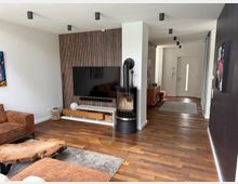 Modern open-plan living room and hallway with hardwood floors, a wall-mounted TV on a vertical wood-slat accent wall, and a freestanding cylindrical black stove on a glass hearth. Recessed ceiling lights line the corridor leading to a white front door with a narrow vertical window.