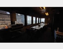 Dunkler Pausenraum mit mehreren rechteckigen Tischen und Stühlen, einer Tischdecke auf den Tischen und einigen Kaffeetassen. Große Fenster bieten einen Blick auf eine industrielle Fabrikumgebung mit Hallen und Maschinen.