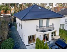 Zweistöckiges modernes Wohnhaus mit weißer Fassade und dunklem Walmdach, Balkon im Obergeschoss, großen Fenstern mit Vorhängen sowie gepflastertem Hofbereich. Umgeben von Bäumen, Sträuchern und Nachbarhäusern in einer grünen Wohngegend.