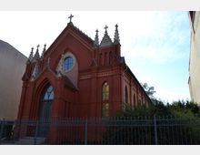 Backstein-Kirchengebäude im neugotischen Stil mit spitzen Türmchen, großen gotischen Fenstern und einem zentralen Eingang mit Spitzbogen. Ein Metallzaun umgibt die Kirche, und im Hintergrund ist ein bewachsener Bereich sichtbar.