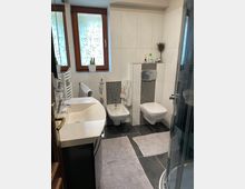 Modernes Badezimmer mit weiß gefliesten Wänden und dunklem Fliesenboden. Ausgestattet mit einem Waschbecken, einem Bidet, einer Wandtoilette, einer verglasten Dusche und einem Fenster mit Milchglas und Pflanzenmuster.