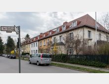 Reihenhäuser mit hellen Fassaden und roten Ziegeldächern in einer Wohnstraße. Im Vordergrund steht ein silberner Van am Straßenrand, umgeben von kahlen Sträuchern und Bäumen, mit einem Straßenschild „Im Gehege“ links im Bild.