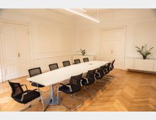 Konferenzraum mit einem langen weißen Tisch, umgeben von schwarzen Netzstühlen auf einem Parkettboden. An den weißen Wänden befinden sich verzierte Paneele, zwei weiße Doppeltüren sowie ein Sideboard mit Zimmerpflanzen.