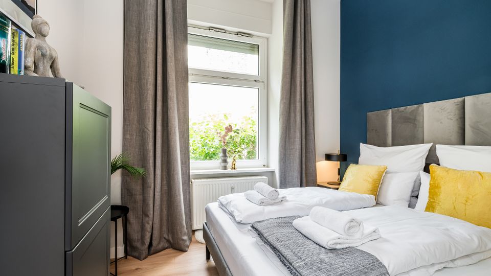 Modernes Schlafzimmer mit Doppelbett, grauem gepolstertem Kopfteil und blauer Akzentwand, weißer Bettwäsche und gelben Kissen. Großes Fenster mit langen grauen Vorhängen über einem Heizkörper sowie ein dunkler Schrank und eine kleine Nachttischlampe.