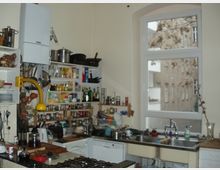 Kleine Küche mit hellen Wänden, einem großen Fenster und offener Regalböden, die mit Geschirr, Gewürzen und Flaschen gefüllt sind. Im Vordergrund befinden sich ein Gasherd, eine Spüle sowie diverse Küchenutensilien.