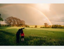 Zwei Personen stehen in einer grünen Wiese und blicken auf einen weiten Horizont, über dem ein klarer Regenbogen vor einem wolkigen Himmel sichtbar ist. Im Hintergrund sind Bäume und Felder zu sehen, die von warmem Licht beleuchtet werden.