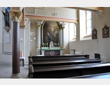 Innenraum einer kleinen Kirche mit Holzbänken, einem verzierten Altar mit Gemälde im Hintergrund und goldenen Akzenten. Rechts befinden sich hohe Fenster, die Tageslicht einfallen lassen, sowie an den Wänden hängende religiöse Bilder.