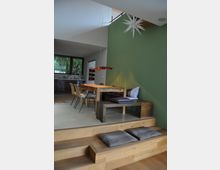 Moderne Wohnküche mit offener Gestaltung; ein Esstisch aus Holz umgeben von Stühlen und einer Sitzbank steht auf einer erhöhten Plattform mit einer grünen Wand im Hintergrund. Im Vordergrund sind Holzstufen mit Kissen, links hinten eine Fensterfront und eine Küche sichtbar.