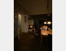 Ein wohnlicher Raum mit einem Esstisch aus Holz und mehreren Stühlen, erhellt durch eine Hängelampe. Im Hintergrund befindet sich ein moderner, weißer Kamin mit einer Feuerstelle, daneben eine geöffnete Tür zu einem angrenzenden Raum.