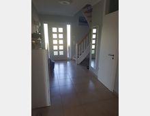 Eingangsbereich eines modernen Hauses mit hellen Wänden, gefliestem Boden und einer weißen Haustür mit Glaseinsätzen. Eine Treppe mit Holzstufen und weißem Geländer führt nach oben, daneben ein großer Wandspiegel und eine geschlossene weiße Tür.