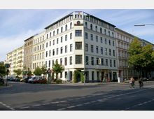 Straßenecke mit mehrstöckigen Wohn- und Geschäftshäusern im Altbau-Stil; das Eckgebäude mit hellem Putz, vielen Fenstern und einem begrünten Balkon. Im Vordergrund eine Kreuzung mit Radfahrern, geparkten Autos und Straßenschildern.