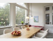 Essbereich mit großem Holztisch, umgeben von cremefarbenen Stühlen und gläsernen Vasen auf der Fensterbank. Große Fenster mit Blick auf einen Garten, zwei hängende Lampen, eine rote Ledercouch und eine weiße Tür runden den Raum ab.