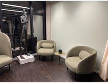 Sitzecke in einem modernen Innenraum mit zwei gepolsterten, olivfarbenen Sesseln, einem kleinen, runden Tisch mit Pflanze und einer großen Skulptur einer abstrakten, stehenden Figur aus Metall auf einem Sockel. Im Hintergrund eine Glaswand mit schwarzem Rahmen, die einen Blick in einen anderen Raum erlaubt.