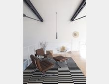 Ein modern eingerichteter Wohnraum mit einer schwarz-weiß gestreiften Teppichfläche, zwei braunen Ledersesseln, passenden Fußhockern und einem kleinen Beistelltisch. Im Hintergrund stehen ein Drahtstuhl mit einem Pelzkissen und eine metallische Hängelampe, während die Decke mit sichtbaren Metallträgern gestaltet ist.