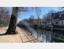 Uferweg entlang eines Kanals mit schmiedeeisernem Geländer, großen Bäumen und einem Fachwerkhaus im Hintergrund; die Szenerie ist von kahlen Bäumen und blauem Himmel geprägt.