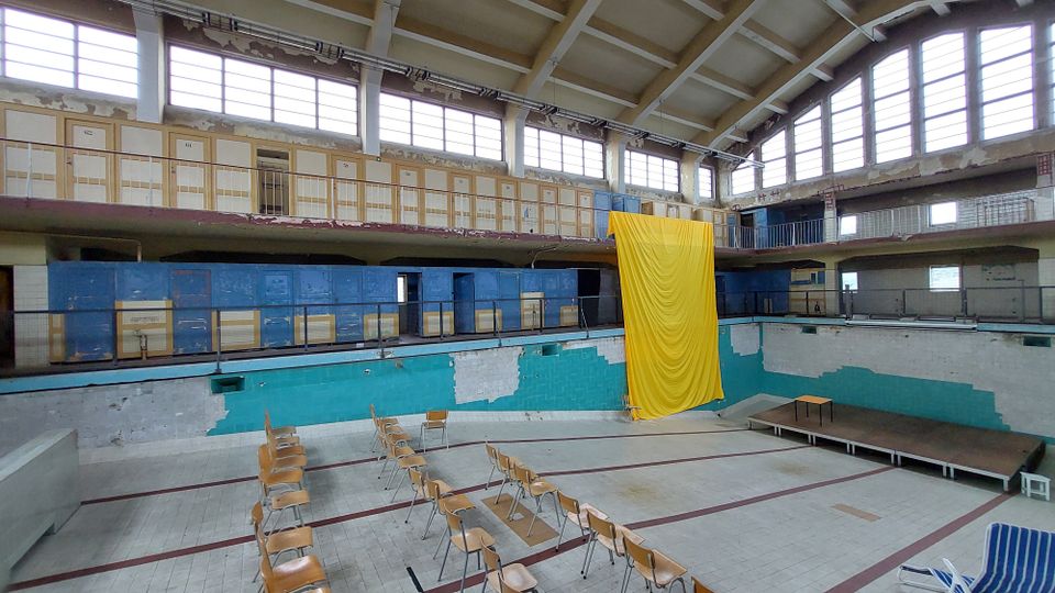 Ein leerstehendes Hallenbad mit leeren Sitzreihen im trockenen Becken, bröckelnden Wänden und einer auffälligen gelben Stoffbahn, die von der oberen Galerie hängt. Die Umgebung zeigt alte Umkleidekabinen über zwei Ebenen und große Fenster, die natürliches Licht einlassen.