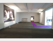 Großer, leerer Galerieraum mit weiß gestrichenen Wänden und einer gewölbten Decke. An der linken Wand hängt eine großformatige Fotografie, während ein lila Teppich den dunklen Boden durchzieht und zu einem beleuchteten kleinen Raum im Hintergrund führt.