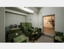 Ein karger Raum mit weißen Wänden aus Ziegeln und Beton, gefüllt mit stapelbaren grünen Metallkisten, von denen einige offen sind und Geräte enthalten. Durch eine geöffnete Tür ist ein weiterer Raum mit technischen Installationen an der Wand sichtbar.