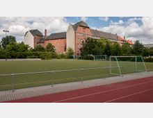 Ein Fußballplatz mit Kunstrasen, umgeben von einem Metallgeländer und einer Laufbahn, im Hintergrund ein großes Backsteingebäude mit Spitzdach und Bäumen. Der Himmel ist teils bewölkt.