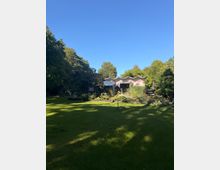 Große Rasenfläche in einem parkähnlichen Garten, umgeben von hohen Bäumen und Beeten mit blühenden Sträuchern. Im Hintergrund steht ein niedriges Gebäude oder Pavillon mit wellenförmigem Dach und einer Terrasse.