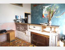 Ein kleiner Innenraum mit einer Theke aus recyceltem Holz und verschiedenen Türfronten, einer Tischlampe und Dekoration aus Zweigen darauf. Die Rückwand ist blau gestrichen, mit einem Poster und einem dekorativen Paddel darauf, während der Boden aus farbigem Holzparkett besteht.