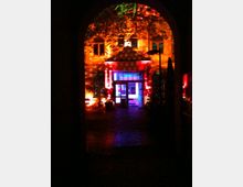 Eine beleuchtete Gebäudefassade in einem Innenhof, mit farbigem Licht in Rot, Blau und Gelb angestrahlt. Im Vordergrund ist ein dunkler Torbogen zu sehen, der den Blick auf den gepflasterten Hof und den Eingang des Gebäudes freigibt.