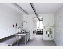 Große, geflieste Industrie- oder Großküche mit weißen Wänden und hellem Boden. Zu sehen sind Edelstahl-Arbeitsflächen, ein Spülbecken mit flexiblem Wasserhahn, an der Wand befestigte Seifenspender sowie ein angrenzender Raum mit einem Fenster.