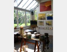 Ein Künstleratelier mit großen Glasfenstern und einer schrägen Glasdecke, die viel Tageslicht hereinlassen. Im Raum stehen ein Tisch mit Farbdosen und Pinseln sowie Staffeleien, während an der Wand gerahmte Bilder und ein großformatiges Gemälde hängen. Im Hintergrund ist ein Garten sichtbar.
