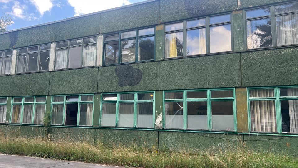 Zweistöckiges, längliches Gebäude mit dunkelgrüner Fassade und Reihen aus großen Fenstern mit grünen Rahmen, teils mit Vorhängen. Vor dem Gebäude verläuft ein Betonweg mit hohem, verwildertem Gras am Rand; darüber ist ein teilweise bewölkter Himmel zu sehen.