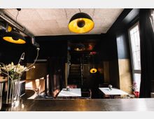 Innenraum eines kleinen Cafés mit dunklen Wänden, großen Hängelampen mit goldener Innenseite und mehreren weißen Tischen mit Stühlen. Im Vordergrund steht eine Theke mit Blumendekoration, und rechts ist ein großes Fenster zu sehen, das Tageslicht hereinlässt.