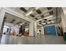 Großes, helles Studio- oder Veranstaltungsloft mit hoher Decke, sichtbaren Trägern, Akustikpaneelen und langen Deckenleuchten über glattem grauem Boden. Im Raum stehen mobile Trennwände, ein blauer Hintergrund, Sitzmöbel und Pflanzen; große Fensterfronten geben den Blick nach draußen frei.
