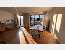 Wohnzimmer mit Holzboden und großen Fenstern, die viel Sonnenlicht hereinlassen. Im Raum befinden sich ein Esstisch mit Stühlen, ein bequemer Sessel, eine Bücherwand und dekorative Elemente wie Pflanzen und Bilder.