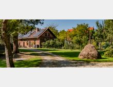 Ziegelbauernhaus mit Solarpanelen auf dem Dach, umgeben von Bäumen und einem gepflegten Garten mit einer gestampften Zufahrtsstraße im Vordergrund; rechts ein großer Heuhaufen auf einer Grünfläche.