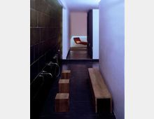 Modern gestalteter Innenraum eines Spa-Bereichs mit dunklen Fliesenboden und Wänden. Links befinden sich mehrere Wandeinbaubäder mit Wasserhähnen, rechts eine lange Holzbank; im Hintergrund ist ein Ruhebereich mit moderner Liege sichtbar.