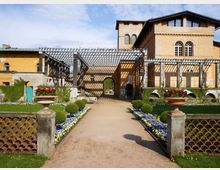 Historisches Gebäude mit hellgelber Fassade und Rundbogenfenstern, umgeben von einem geordneten Garten mit Blumenbeeten, Buchsbaumkugeln und Terrakotta-Pflanzgefäßen. Eine überdachte Pergola mit Holzstreben führt zu einem Torbogen am Ende des Gehwegs.