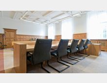 Konferenzraum mit einem großen rechteckigen Holztisch, umgeben von mehreren gepolsterten Stühlen mit Metallfüßen. Die Wände sind mit hellen Holzpaneelen verkleidet, es gibt große Fenster mit vertikalen Jalousien, und die Decke hat eine dekorative Kassettengestaltung.