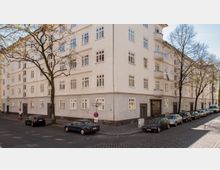 Eine Straßenecke mit einem mehrstöckigen, hell verputzten Wohngebäude im Stil der frühen 20. Jahrhunderts, das zahlreiche rechteckige Fenster und Balkone aufweist. Die Umgebung zeigt eine gepflasterte Straße mit geparkten Autos und spärlich belaubten Bäumen.