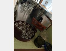 Eine Theke mit Zapfhähnen im Hintergrund, daneben Biergläser und kleine Gläser auf einem Holztablett. Im Vordergrund steht ein Holzschild mit der Aufschrift „Stammtisch“.