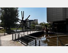 Ein moderner Platz mit einer Fußgängerbrücke über ein Wasserbecken, gesäumt von Geländern. Links steht eine große abstrakte Metallskulptur, umgeben von Bäumen. Rechts ist ein Gebäude mit der Aufschrift "Viet Bowl", daneben stehen große Pflanzkübel mit Palmen.