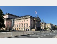 Historisches Gebäude mit klassizistischer Architektur, rosa Fassade, einem dreieckigen Giebel mit Statuen und Säulen am Eingang. Im Vordergrund sind eine gepflasterte Straße und einige Verkehrsschilder zu sehen.