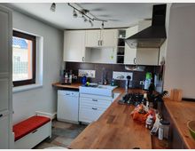 Kleine, helle Küche mit weißen Wandschränken, Holzarbeitsplatten und einer Spüle unter einem Fenster. Es gibt eine Dunstabzugshaube über einem Kochfeld, eine Spülmaschine, und diverse Küchenutensilien sind auf der Arbeitsfläche verteilt.