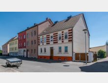 Straßenansicht einer Reihe mehrgeschossiger Wohnhäuser mit unterschiedlichen Fassadenfarben, darunter Gelb, Rot und Beige. Im Vordergrund steht ein Anhänger auf der asphaltierten Straße, und rechts ist ein Gartenzaun sowie eine Straßenlaterne sichtbar.