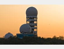 Ruine einer ehemaligen Abhörstation mit mehreren geodätischen Kuppeln auf einem Turm, umgeben von Bäumen, vor einem orangefarbenen Himmel bei Sonnenuntergang. Ein kleines Flugzeug ist am Horizont sichtbar.