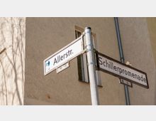 Straßenschilder an einer Kreuzung mit den Aufschriften „Allerstr.“ und „Schillerpromenade“, befestigt an einem Metallpfosten, im Hintergrund eine Hausfassade mit Fenster.