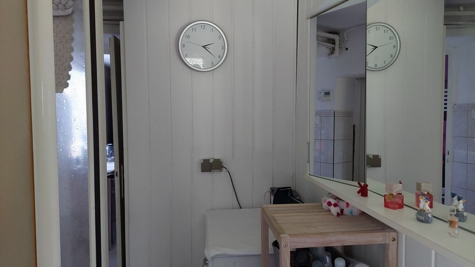 Schmaler Innenraum mit hellen Holzpaneel-Wänden, einer runden Wanduhr und einem großen Wandspiegel über einer Ablage. Daneben stehen ein kleiner Holztisch und ein Hocker; links ist eine Glastür zu einem angrenzenden Raum sichtbar.