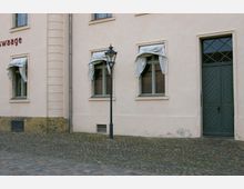 Fassade eines historischen Gebäudes mit hellbeigem Putz, drei Fenstern mit hochgebundenen Vorhängen, einer grün gestrichenen Doppeltür mit Glasoberlichtern und einer schwarzen Laterne auf einem gepflasterten Vorplatz.