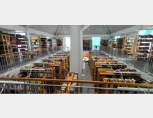 Bibliothek mit mehreren Etagen und offenen Regalen voller Bücher. Ein zentraler Pfeiler teilt den Raum, und Geländer begrenzen die Galerieebene, während durch große Fenster Tageslicht einfällt.