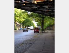 Straßenansicht mit Kopfsteinpflaster, vorbei an grünen Bäumen und geparkten Autos; im Vordergrund verläuft ein Bürgersteig, und oben ragt eine Stahlbrückenkonstruktion hinein.