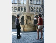 Ein Innenhof mit gepflastertem Boden, einem verzierten Steinbrunnen mit zentraler Säule und Reliefs, umgeben von einer Fassade mit Arkadenbögen und Fensterreihen. Zwei Personen stehen in der Nähe des Brunnens und betrachten ihn.