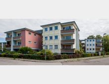 Wohnanlage mit mehrstöckigen Wohnhäusern, die helle Fassaden in Beige und Rosa sowie Balkone mit braunen Geländern aufweisen. Im Vordergrund befinden sich gepflegte Hecken und Gehwege, daneben ein Straßenschild und eine Straßenlaterne.